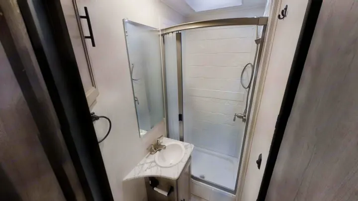 View inside an RV rental bathroom, showing a small vanity with sink, mirror, and curved shower.. Coachmen Freelander 2021