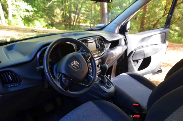 Inside an RV rental van: driver's view of dashboard, steering wheel (Ram logo), and manual transmission. Open door shows outdoor scenery.. Dodge Ram Promaster City 2016