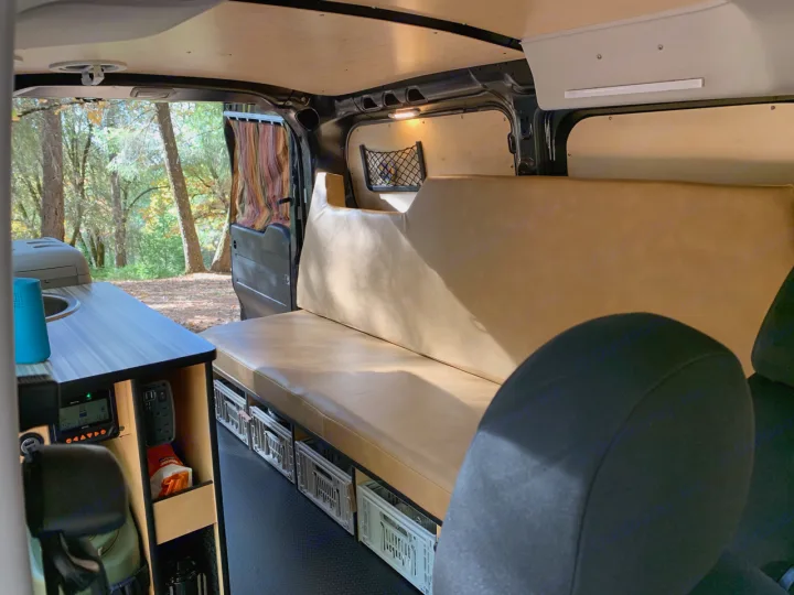 Inside an RV rental camper van: a cozy bench bed, kitchen, storage crates, and forest views from the open door.. Dodge Ram Promaster City 2016