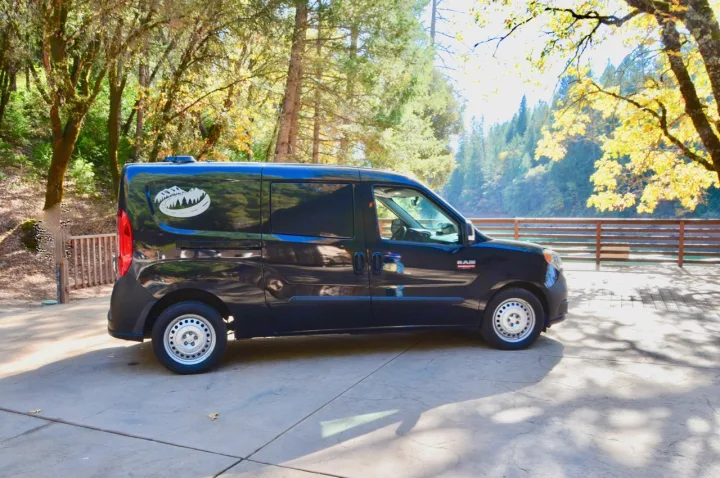 A black compact van, an RV rental, parked outdoors by a river, surrounded by vibrant forest. Ideal for travel adventures.. Dodge Ram Promaster City 2016