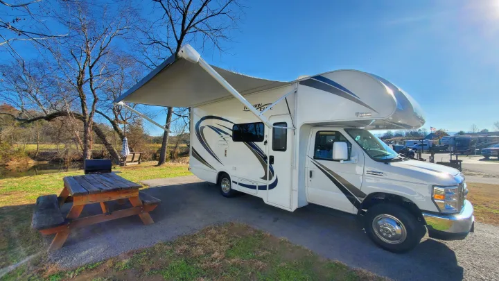 White rv rental motorhome with awning extended at a sunny campsite by a river, next to a picnic table. Ideal for outdoor travel.. Thor Freedom Elite 2019