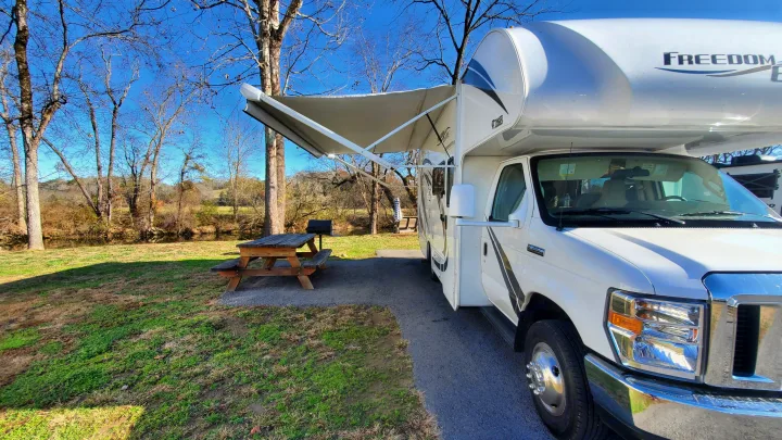 Class C RV rental with awning extended at a riverside campsite. Includes picnic table & grill under clear sky.. Thor Freedom Elite 2019