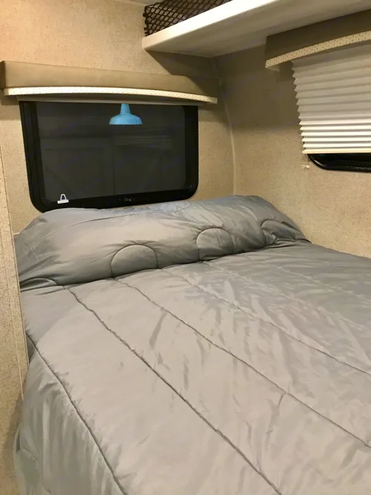 Comfortable gray bed in a compact RV rental interior with windows, overhead storage, and light fixture reflection.. Other Other 2021