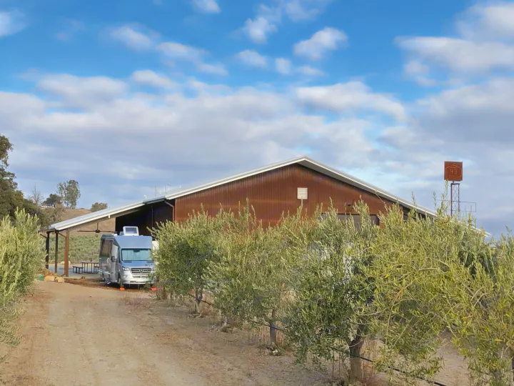 A silver Mercedes Sprinter rv rental parked beside a brown farm building, with olive trees lining a dirt road under a cloudy sky.. Airstream Atlas 2021