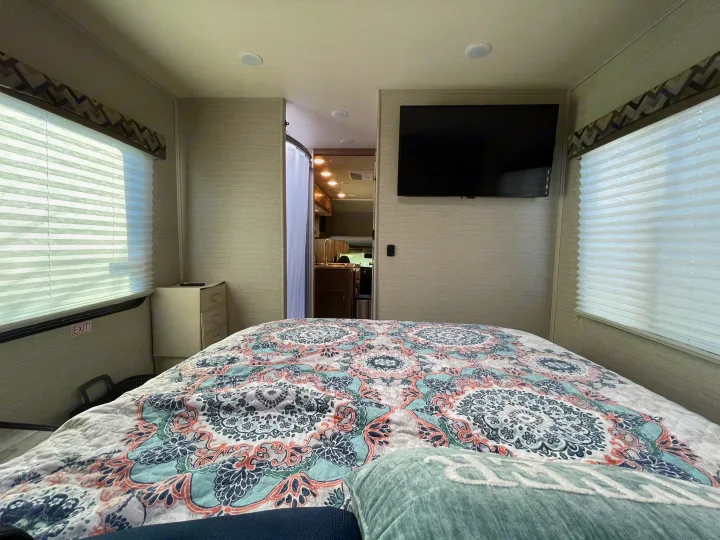 Bright RV rental bedroom: patterned bed, wall TV, windows. Exit sign visible. Leads to kitchen.. Jayco Redhawk 2020