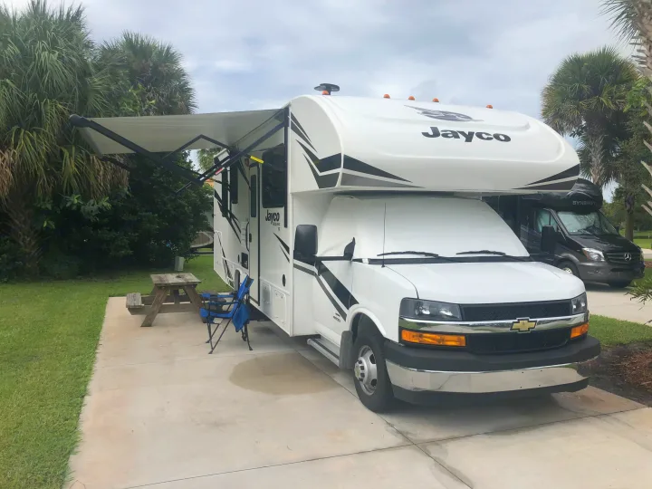 This Jayco RV rental is set up at a scenic campsite with an extended awning, picnic table, and chairs, ready for outdoor living.. Jayco Redhawk 2020