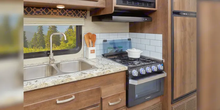 RV rental kitchen: compact interior with double sink, stove, oven, fridge. Window offers a scenic view of trees & mountains.. Jayco Redhawk 2020