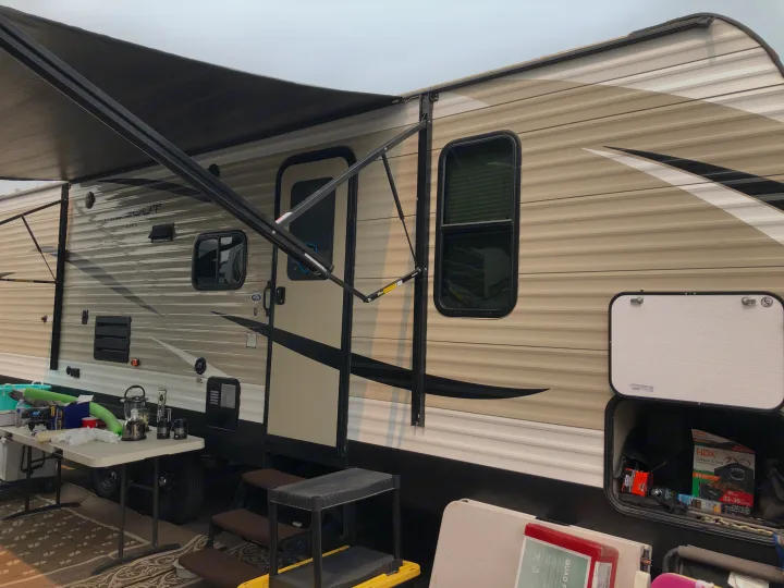 This rv rental is set up at a campsite with an extended awning, outdoor table, and open storage compartment.. Keystone Hideout 2018