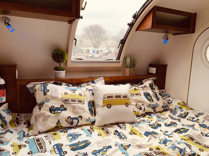 Cozy RV rental interior: patterned bed with vehicle theme, accent pillow, and window view of a USPS truck.. nuCamp T@G 2019