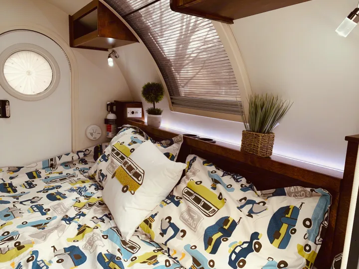 Inside a cozy RV rental: a bed with playful car bedding, a round door, and ambient lighting.. nuCamp T@G 2019