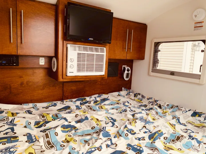 Cozy RV rental interior with a bed, TV, AC, and wooden cabinets. Playful car-themed bedding adds a fun touch.. nuCamp T@G 2019