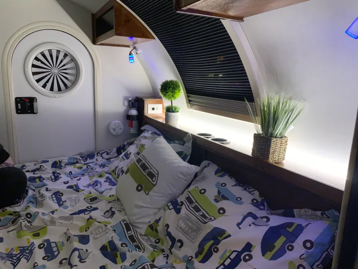 Cozy RV rental sleeping area: bed with car-themed bedding, circular window, blue accent & LED lighting, safety gear.. nuCamp T@G 2019