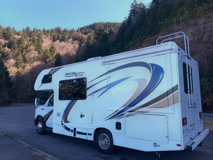 White Freedom Elite RV rental parked by a forested mountain under clear skies, ideal for outdoor adventures.. Thor Freedom Elite 2019