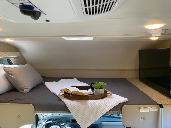 Cozy RV rental cab-over bunk features a bed, TV, and thoughtful comforts like a book and plant.. Thor Freedom Elite 2019