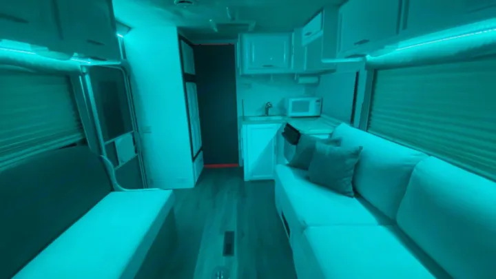 Bright teal-lit RV rental interior featuring a living area with facing sofas, a compact kitchen, and wood flooring.. Jayco Greyhawk 2002