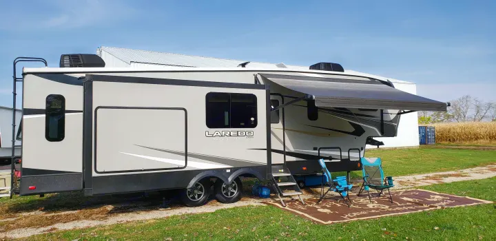 A grey fifth-wheel RV rental with an extended awning, chairs, and mat set up on a grassy patch.. Keystone RV Laredo 2020