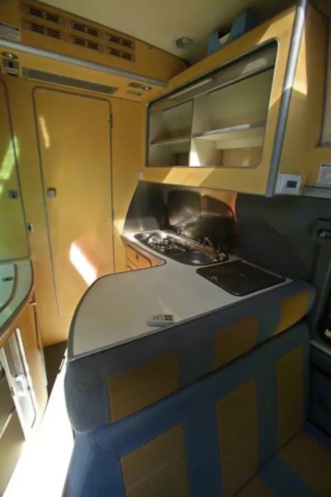 Compact RV rental interior showcasing a functional kitchen with sink/stove, overhead storage, and comfortable, striped seating.. Airstream Westfalia 2005