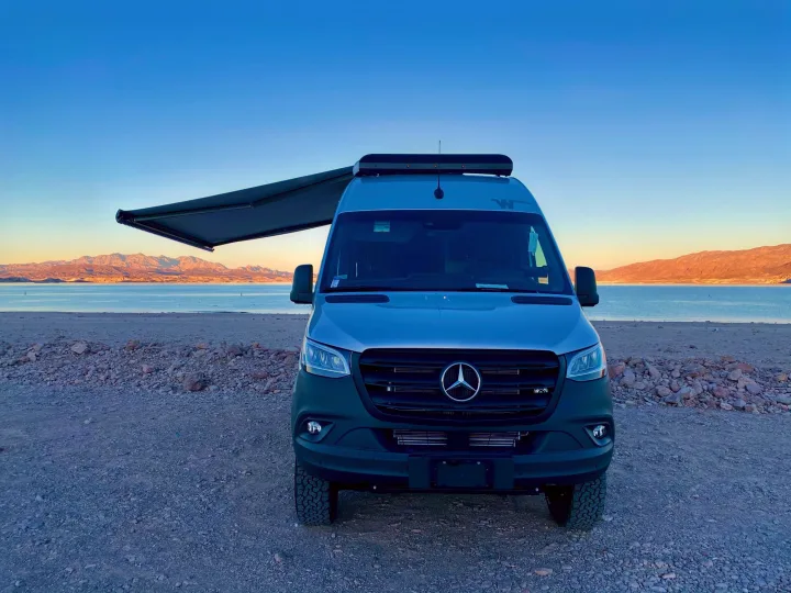 Front of an RV rental van with awning extended, parked on gravel by a calm lake & distant mountains.. Winnebago Revel 2021