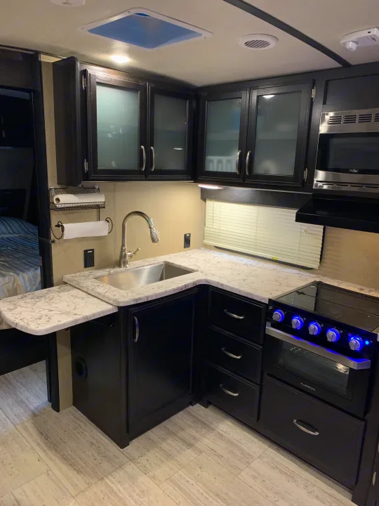 An rv rental kitchen featuring dark cabinets, light counters, a sink, stove with blue lit knobs, and an overhead microwave.. Grand Design Imagine 2019