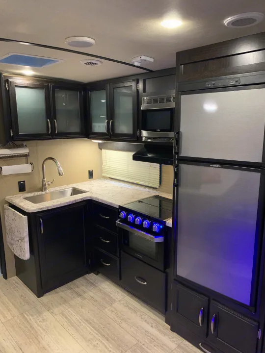 Well-equipped RV rental kitchen featuring dark cabinets, light counters, stainless microwave, Dometic fridge, and Furrion oven with blue lights.. Grand Design Imagine 2019
