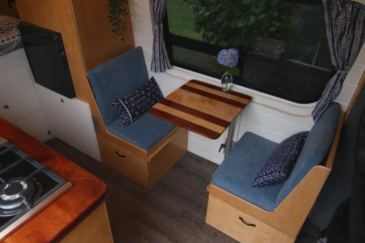 Cozy RV rental interior showcasing a compact dining area with blue benches, a wooden table, and an integrated kitchen.. Mercedes-Benz Sprinter 2012