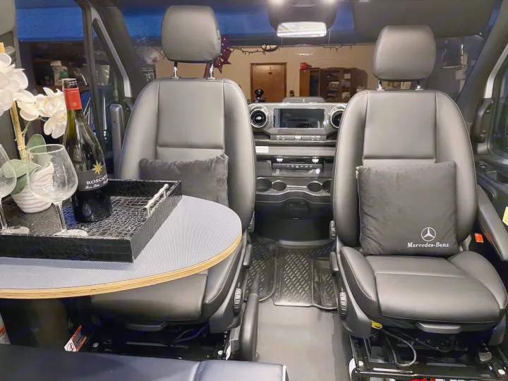 Luxury RV rental interior: black Mercedes-Benz front seats, a dashboard, and a side table with wine and glasses.. Winnebago Revel 2021
