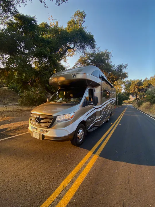 A golden Mercedes-Benz RV rental parked on a sunlit, tree-lined road, ready for a scenic journey.. Winnebago Navion 2021