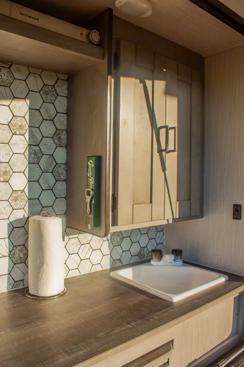Compact RV rental kitchen features a sink, hexagonal backsplash, cabinetry, and paper towel holder, bathed in sunlight.. Heartland M-33 2021