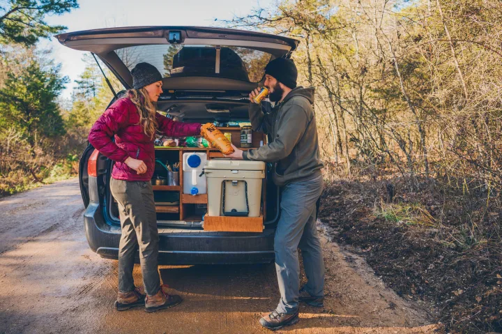 Couple shares snacks from their organized car-camping setup, ideal for a remote **rv rental** adventure on a dirt road.. Toyota Other 2007