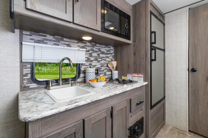 Compact RV rental kitchen with a sink, cooktop, microwave, and fridge, all in modern wood-grain cabinetry.. Gulf Stream Amerilite 2021