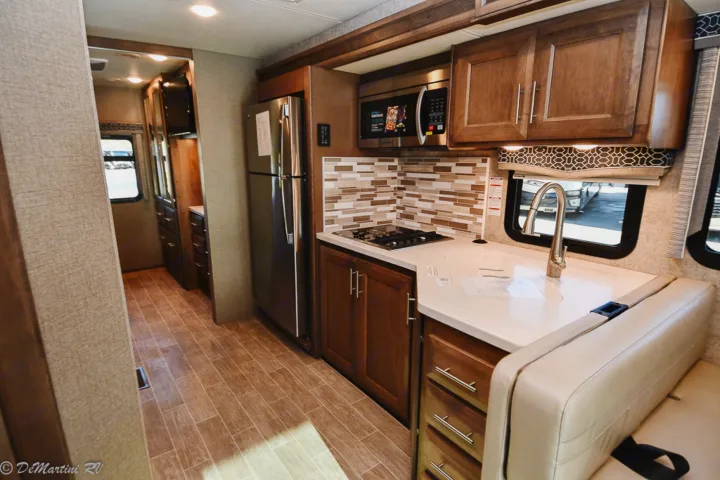 Modern RV rental interior: kitchen with microwave, stove, sink, fridge, and dinette seating. Hallway and wood-look flooring visible.. Thor Motor Coach Other 2021