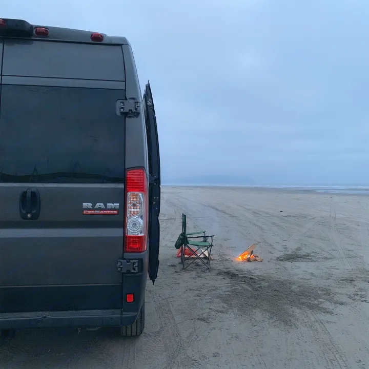 Dark grey RV rental van, a Ram ProMaster, parked on a vast sandy beach at dusk. A camping chair and glowing campfire are beside it.. Dodge Ram 1500 2019