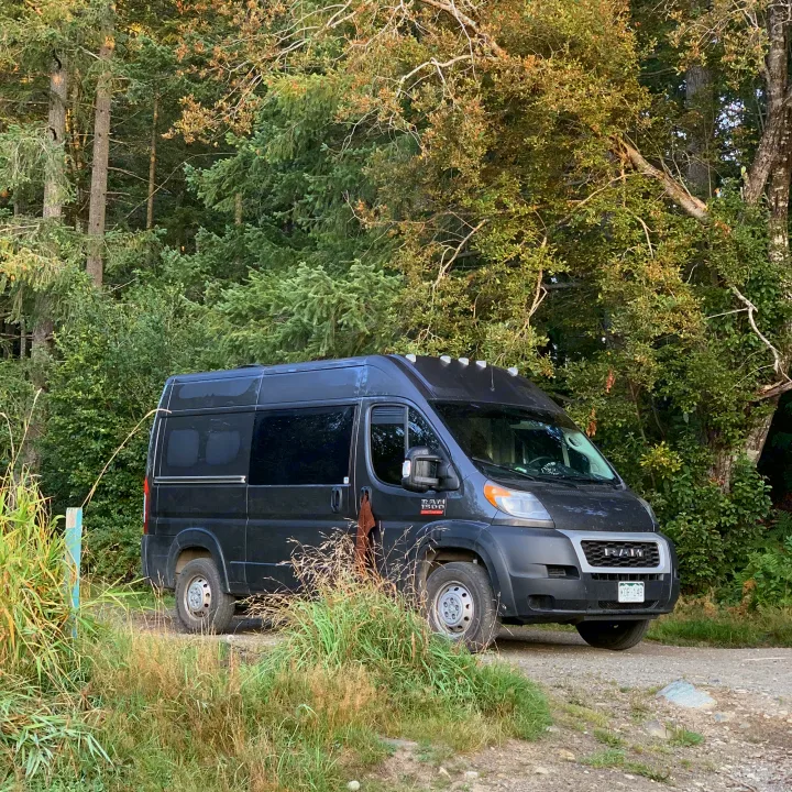 Dark grey RV rental camper van, a Ram ProMaster, parked on a gravel path near a dense forest.. Dodge Ram 1500 2019