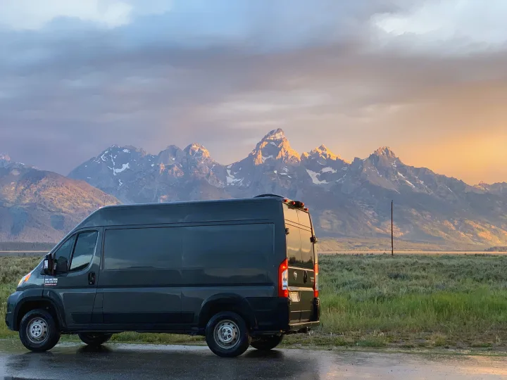 An RV rental camper van faces majestic Grand Teton mountains at sunset. Snow-capped peaks glow under a dramatic sky.. Dodge Ram 1500 2019