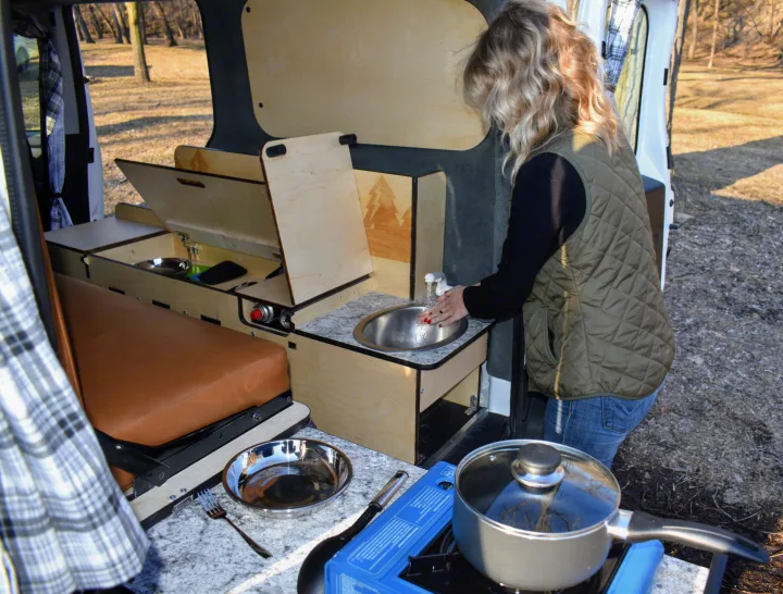 An RV rental provides a compact outdoor kitchen with a sink & cooking area. Woman washes hands.. Dodge Promaster City 2020