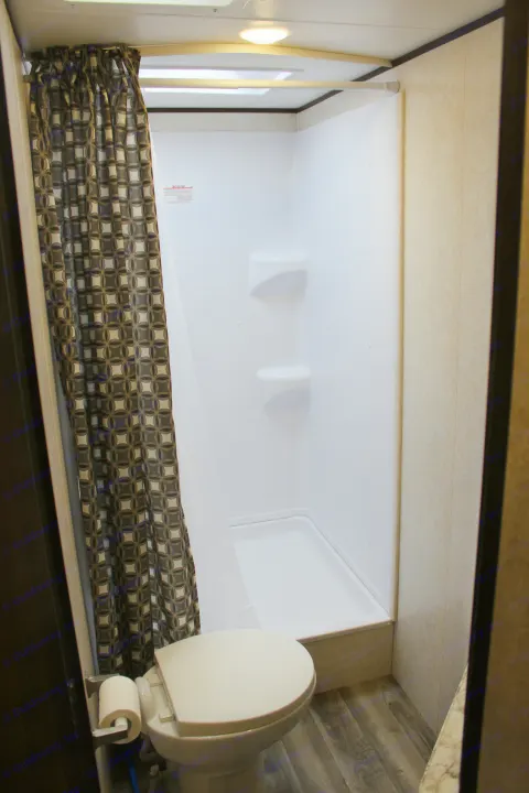 Compact RV rental bathroom featuring a toilet, patterned shower curtain, and a clean shower stall with built-in shelves.. Jayco Jay Flight SLX Rocky Mtn Edition 2019