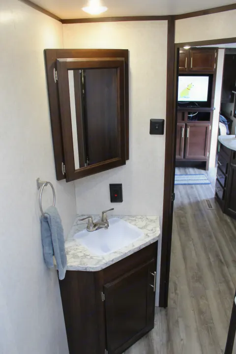 RV rental bathroom features a compact vanity, white sink, mirrored cabinet, and towel ring. A peek into the living area is visible.. Jayco Jay Flight SLX Rocky Mtn Edition 2019