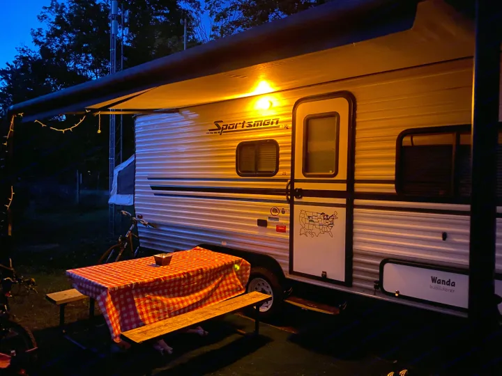 Cozy evening camping with an RV rental! A lit awning illuminates a picnic table with a red checkered cloth, ideal for outdoor dining.. K-Z Manufacturing Sportsman Classic 2015