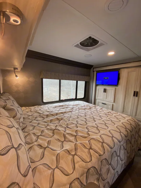 Inside an RV rental bedroom: cozy bed with patterned linens, TV, cabinetry, and window view.. Forest River Forester 2021
