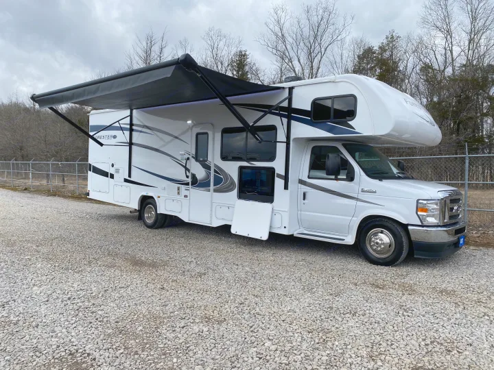 Explore comfortably in this white Class C rv rental! Features an extended awning, outdoor TV, parked on gravel.. Forest River Forester 2021