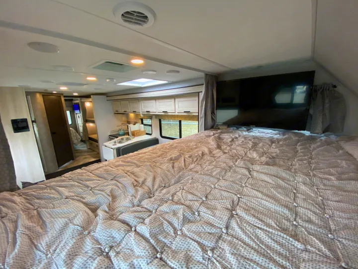 Interior of an RV rental: a cozy bedroom with a patterned bed and TV. A compact kitchen is visible in the background.. Forest River Forester 2021