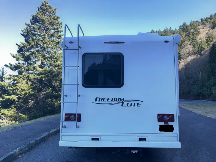 Rear of a white Freedom Elite RV rental with ladder & window, parked by trees under clear sky.. Thor Freedom Elite 2019