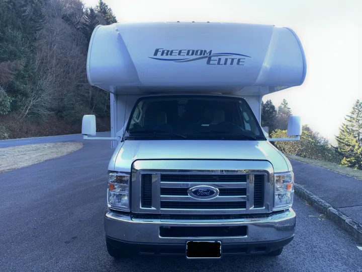 Explore nature with this white Ford Freedom Elite RV rental! Front view of a spacious motorhome parked among trees.. Thor Freedom Elite 2019