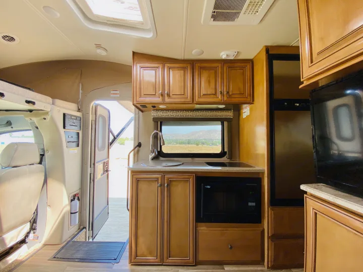 This RV rental interior shows a compact kitchen with a sink, cooktop, microwave, and wood cabinets. An open door provides an outdoor view.. Thor Motor Coach Other 2016