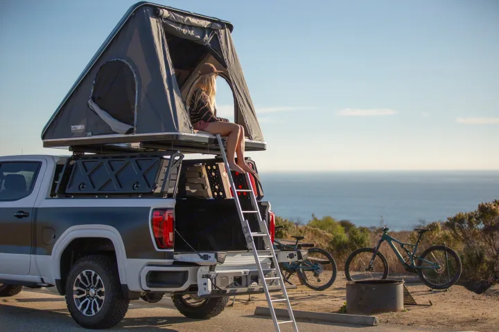 Scenic RV rental alternative: a pickup truck with a rooftop tent overlooking the ocean. A woman relaxes inside, bikes nearby for adventure.. Other Other 2021
