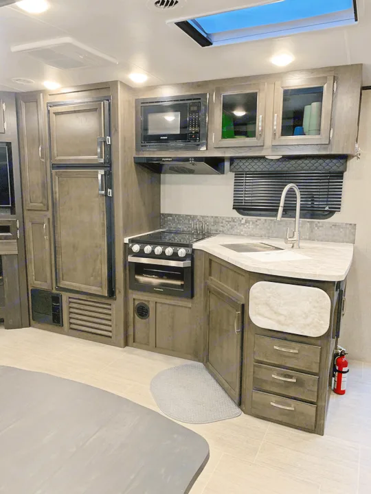 A modern RV rental kitchen features dark wood cabinets, a full refrigerator, stove, microwave, and sink, under bright ceiling lights.. Jayco White Hawk 2021