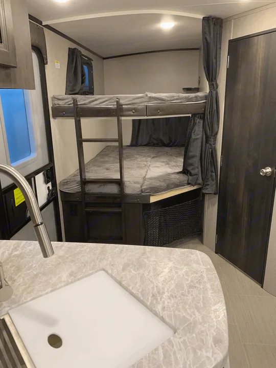 RV rental interior showcasing bunk beds with ladder & privacy curtains, a kitchen sink, and a nearby entry door. Efficient use of space.. Jayco White Hawk 2021
