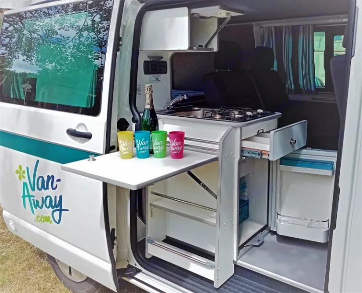 An RV rental camper van's open side door reveals a compact kitchen with stove, sink, and a pull-out table.. Volkswagen Transporter T6.1 2021