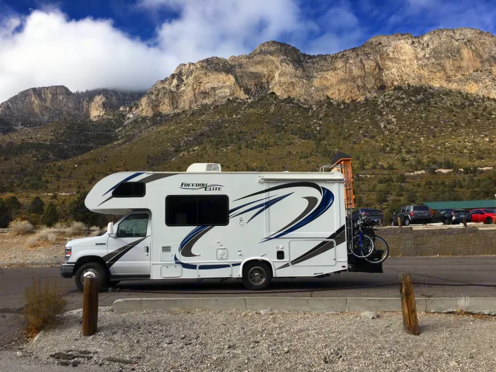 A white Freedom Elite RV rental with a bike rack is parked against a backdrop of rugged mountains and blue sky.. Thor Freedom Elite 2018