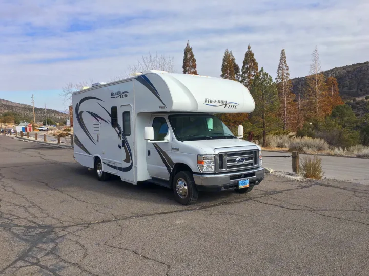 A white Freedom Elite Class C RV is parked on a cracked asphalt road. Mountains and trees create a scenic backdrop for your rv rental travel.. Thor Freedom Elite 2018